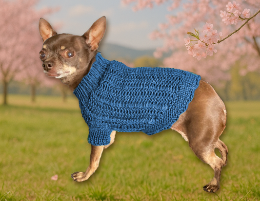 A small brown chihuahua sits on green grass beneath blooming cherry blossom trees, with rolling hills in the background under a clear spring sky